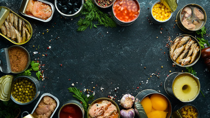 Canned vegetables, beans, fish and fruits in tin cans on black stone background. Food stocks.