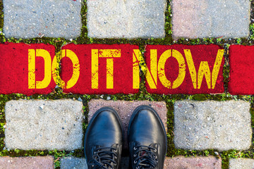 Zwei schwarze Damenstiefel vor einer roten Linie mit dem gelben Text DO IT NOW aus der...
