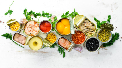 Food background in tin cans. Canned vegetables, beans, fish and fruits on a white wooden background. Top view.