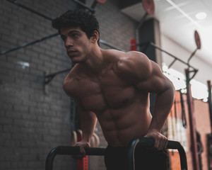 Young, shirtless, muscular athlete doing some dips in parallel bars in the gym. Professional calisthenics athlete working out - obrazy, fototapety, plakaty