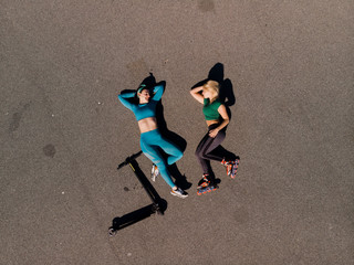 Two happy female friends with roller skates and electric scooter lie on pavement