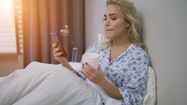 Beautiful young woman browsing smartphone and holding mug of hot beverage sitting on bed