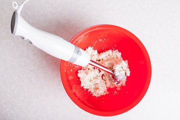Submersible blender with a light handle and a metal nozzle in a red bowl with mixed curd and sugar for making cheesecakes for breakfast close-up isolated on a white background.