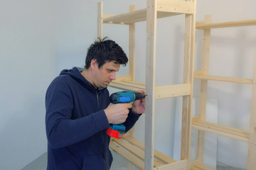 Portrait of man screwing parts with bolts using electric screwdriver collecting rack furniture. Assembling wooden bookcase on floor at home house. Moving at new place. Guy making renovation.