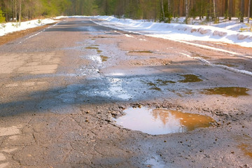 Destroyed broken asphalt country road with holes, melting snow and puddles early spring. Old village road after winter. Cracked ruined bad rural asphalt in forest, national park.