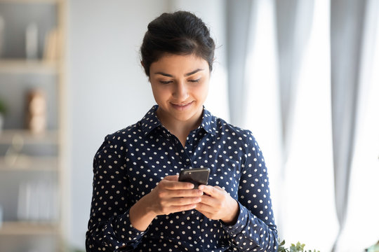 Head shot pleasant millennial indian girl holding smartphone, reading good news sms, using mobile applications at home. Happy young hindu student woman enjoying spending time in social network.