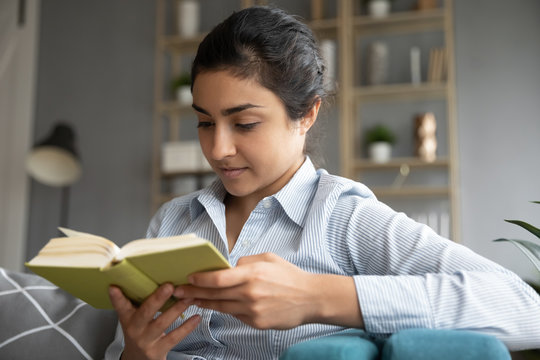 Head shot close up happy focused pretty young indian woman enjoying spending free weekend leisure time, reading paper book bestseller novel story, sitting on comfortable sofa alone in living room.