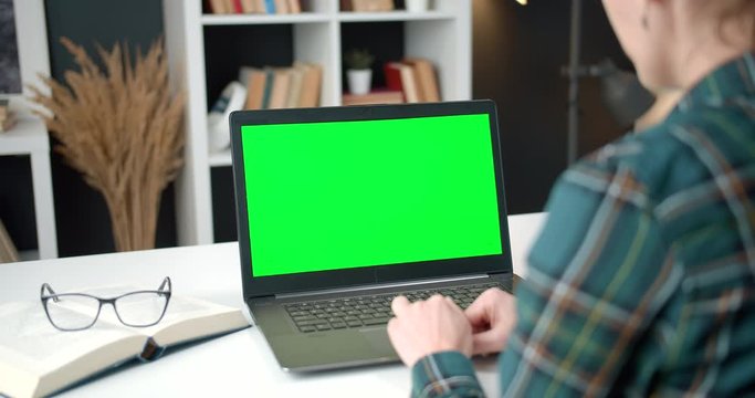 Mature woman in casual clothing sitting at table and working on personal laptop with empty green screen. Female with dark hair doing his job remotely at home.
