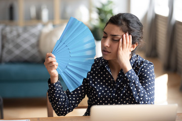Head shot young exhausted indian woman waving paper fan, suffering from high temperature at home....