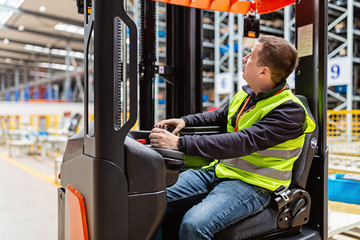 Storehouse employee in uniform working on forklift in modern automatic warehouse. Boxes are on the...