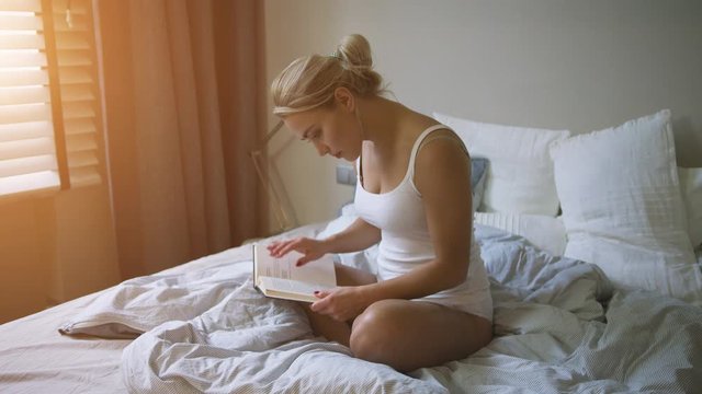 Lovely young woman in white tank top and panties sitting on comfortable bed and reading