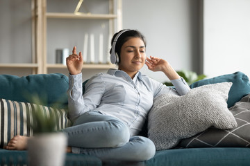 Full length young happy indian girl resting on cozy sofa, listening to energetic music in headphones. Smiling peaceful carefree young woman enjoying spending free time with favorite audio tracks.