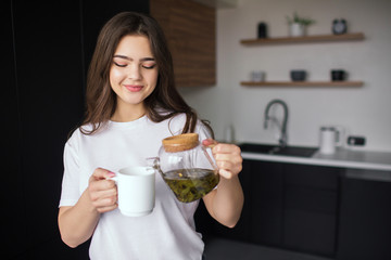 Young woman in kitchen during quarantine. Girl in casual look and white shirt pouring some tea from teapot into white cup. Going to drink it.