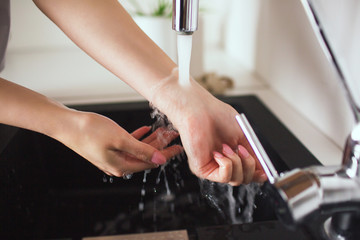 Young woman in kitchen during qiarantine. Cut view of gorl's hands holding under water jet come out of tap. Washing or warmiing up hands and wrist.