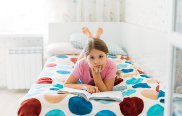 Cute tween girl in pink t-shirt reading book lying on bed in bright room at home