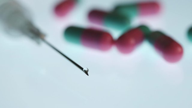 Tip of Syringe with Drop Coming Out From Needle, Capsules on White Background. 4k