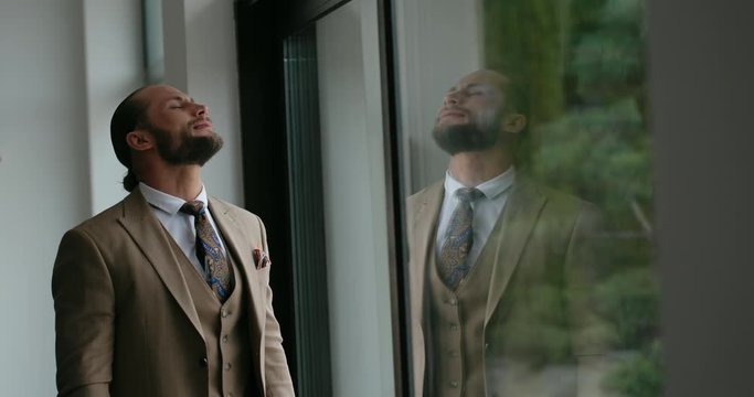 Bearded male in elegant suit tilting head back in pain then feeling relief while standing near window inside modern building