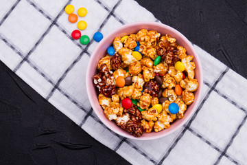 Crispy popcorn and chocolate candies mix on napkin against brown wooden background. Checkered white napkin.