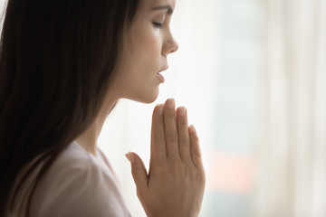 Close up side view of religious young woman hold hands in prayer asking God for good luck or fate, superstitious female believer pray at home, believe in future, faith, religion, superstition concept