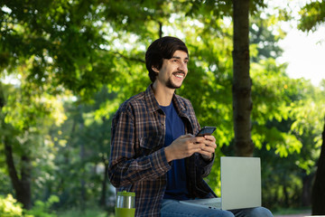 Young freelancer smiling sitting with a laptop in a park and holding smartphone.