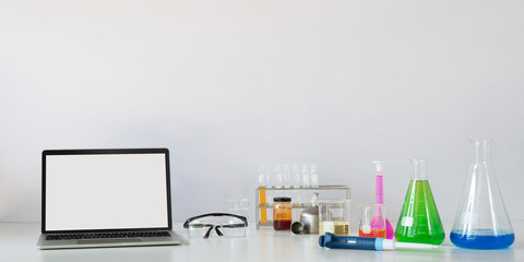 Computer laptop with white blank screen putting on white working desk that surrounded by chemistry glass ware over laboratory white wall as background.