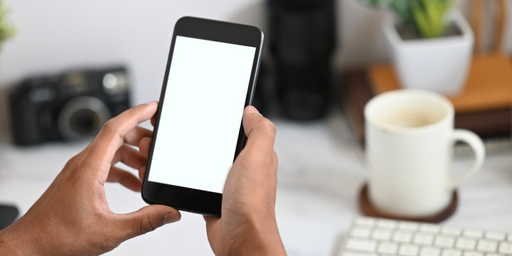 Cropped image of hands holding a cropped black smartphone with white blank screen over working desk that surrounded by office equipment as background.