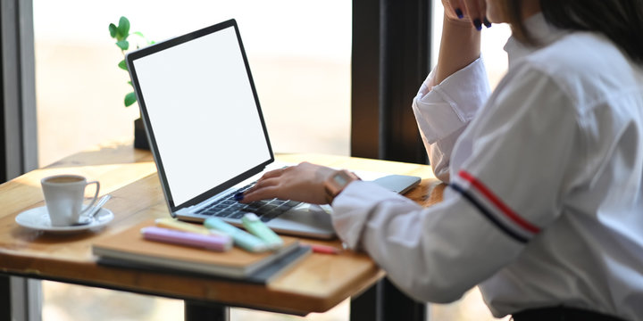 Cropped image of beautiful woman in white shirt keep hand on chin and  typing on computer laptop with white blank screen while sitting at the wooden working desk over office glass wall as background.