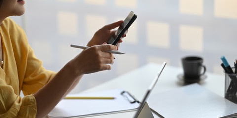 Photo of stylish woman holding a smartphone and stylus pen in hands while sitting in front her computer tablet with keyboard case that putting on white working desk over modern office as background.