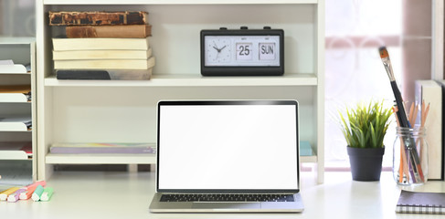 Photo of computer laptop with white blank screen putting on white student desk and surrounded by clock, books, potted plant, pencils in glass vase, notebook and paint brush.