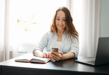 Adult smiling brunette woman doing notes in daily book and using the mobile phone with opened...