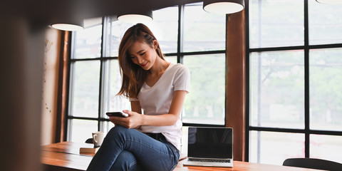 Photo of beautiful stylish woman using a smartphone while sitting with crossed leg on the wooden long table over comfortable living room windows as background.