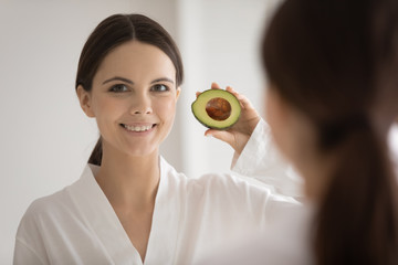 Portrait of smiling young Caucasian woman look in mirror holding piece of avocado recommend natural beauty products for healthy glowing skin, happy female prefer organic cosmetics, skincare concept