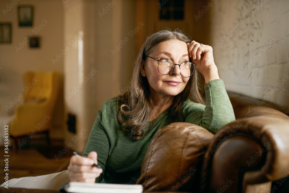 Wall mural Stylish inspired middle aged woman writer wearing glasses sitting in leather couch with pen making notes. Attractive mature female artist sketching or teacher preparing tasks for online lessons