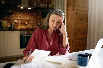 Indoor image of frustrated female pensioner managing finances at home, sitting at desk with...