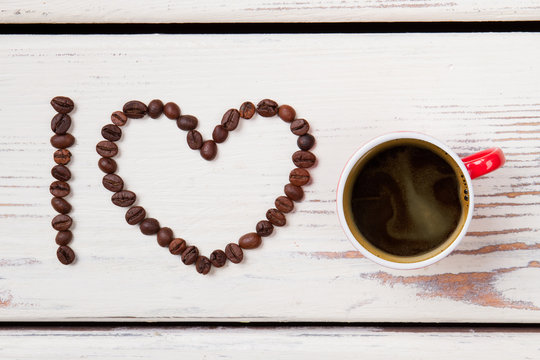 Red cup of coffee and beans arranged in a heart shape. I love coffee. White wood on background.