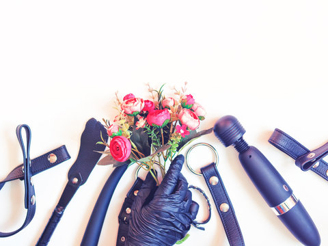 Various sex toys are on a white background. A male hand in a black latex glove holds a bouquet of flowers. Nearby are sex toys (dildo, harness, vibrator and others). Flat lay, copy space.