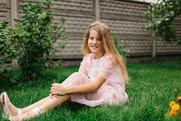 Naklejka na ściany i meble beautiful blue-eyed girl with long blond hair. little girl in a pink flamingo dress. summer bright, emotional photo. girl model with long hair sits on green grass