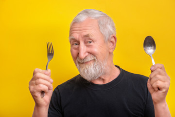 Senior man smile and holds spoon and fork and over yellow background with copy space. Senior man...