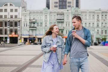 Romantic and happy caucasian couple in casual clothes walking together through the streets. Love, relationships, romance, happiness concept. Man and woman with disposable cups of coffee in the city.
