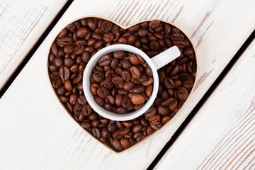 Cup and heart-shaped form filled with coffee grains. Top view flat lay. White wood on background.