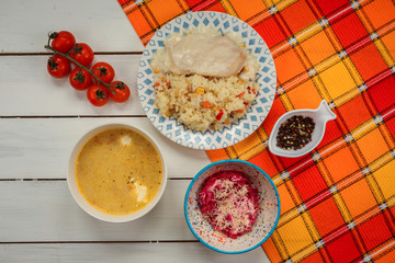 Tasty and appetizing lunch set consists of soup with carrot, beet salad and rice with vegetables and chicken steak. White wooden table and orange table. Food delivery set. 