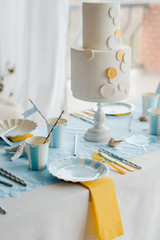 Festive table setting in sea style. Elegant plates, paper cups, straws and cutlery with blue and yellow textile. Paper boats with sweets. Birthday or baby shower boy.