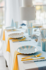 Interior decoration and table setting with white dishes, yellow cutlery, bluel tablecloth. Birthday party decoration. Selective focus.