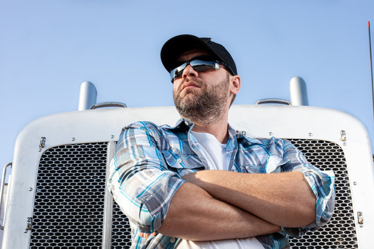 Confident semi truck driver wearing plaid shirt and black baseball cap  stands with arms crossed in front of big rig. Portrait of professional looking trucker. Blue collar jobs in transportation. 