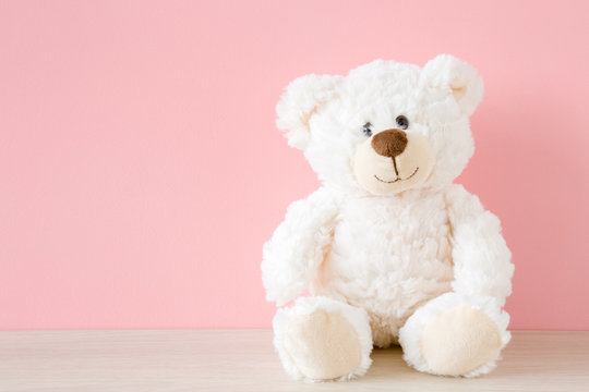 Naklejki Smiling white teddy bear sitting on table at pastel pink wall background. Kids best friend. Front view. Close up.