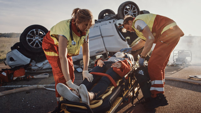 On the Car Crash Traffic Accident Scene: Paramedics Saving Life of a Female Victim who is Lying on Stretchers. They Apply Oxygen Mask and Give First Aid. In Background Rollover Vehicle