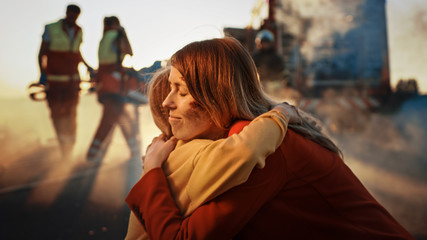 Injured Mother and Young Daughter Reunite After Terrible Car Crash Traffic Accident, They Hug Happily. In the Background Through Smoke and Fire, Courageous Paramedics and Firefighters Save Lives - obrazy, fototapety, plakaty
