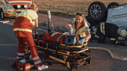 On the Car Crash Traffic Accident Scene: Paramedics Saving Life of a Female Victim who is Lying on Stretchers. They Apply Oxygen Mask, Do Cardiopulmonary Resuscitation / CPR and Perform First Aid © Gorodenkoff