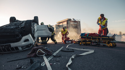 Paramedics and Firefighters Arrive On the Car Crash Traffic Accident Scene. Professionals Rescue Injured Victim Trapped in Rollover Vehicle by Extricating Them, giving First Aid and Extinguishing Fire © Gorodenkoff