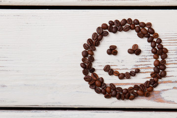 Smiling emoticon made of coffee beans. White wooden surface.